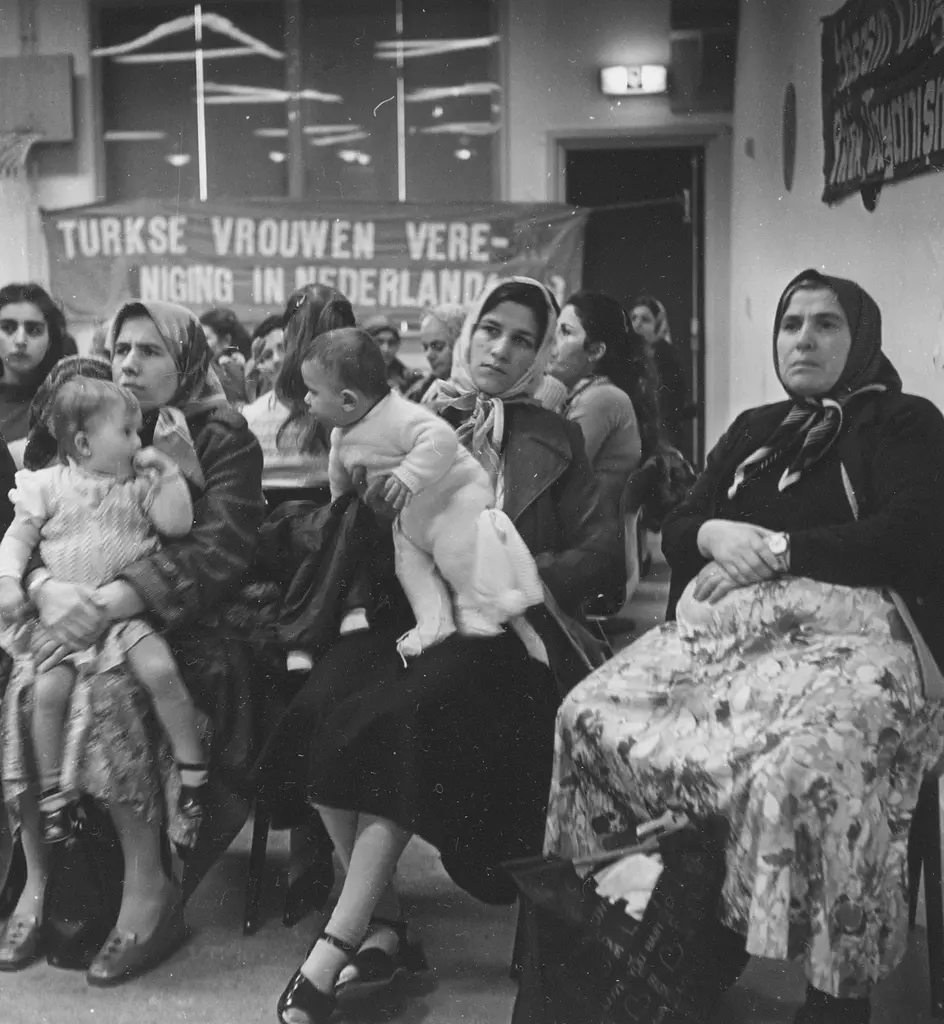 Groep Turkse vrouwen zit in een buurthuis te luisteren. Een paar moeders houden een kind op schoot.