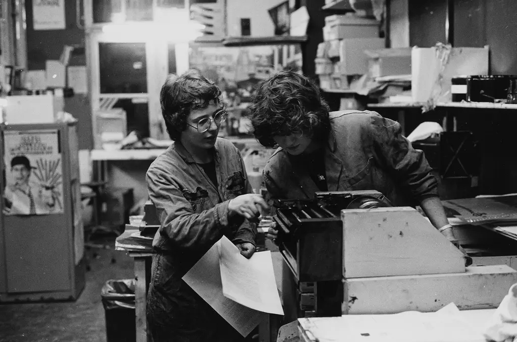 Twee vrouwen aan het werk in een drukkerij. Ze hebben vellen papier in hun handen en kijken naar een drukpers of printer. Zwart-witte foto.