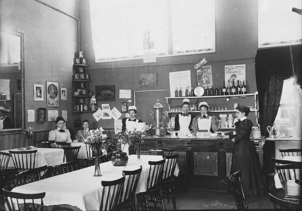 Groep vrouwen in een restaurant in werkkleding. Ze staan deels achter de bar en achter lange gedekte tafels met witte tafelkleden.