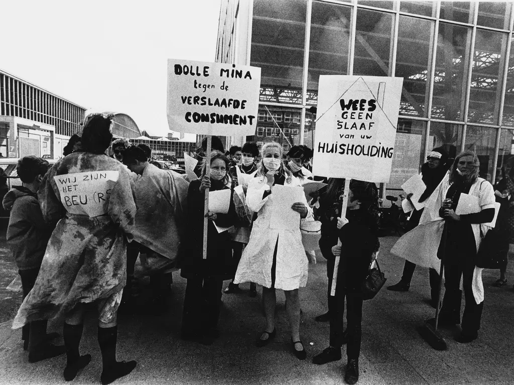 Actiegroep Dolle Mina protesteert bij de Huishoudbeurs. Ze houden borden vast met teksten als "Dolle Mina tegen de verslaafde consument" en "wees geen slaaf van uw huishouding" en "wij zijn het beu(rs)"