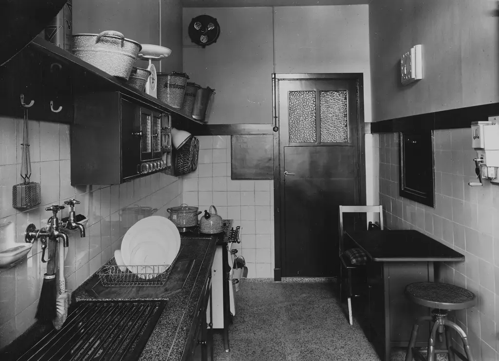 Modelkeuken van de Nederlandse Vereniging van Huisvrouwen. Zwart-witfoto van een ouderwetse keuken met een klein tafeltje met een barkruk ernaast.