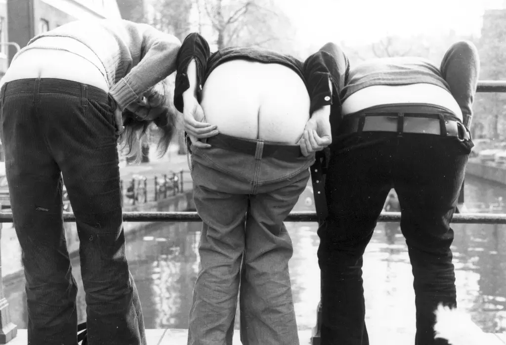 Zwart-wit foto van de drie vrouwen van Paarse September die op een brug staan en hun broek een stuk naar beneden hebben getrokken om hun achterste te laten zien