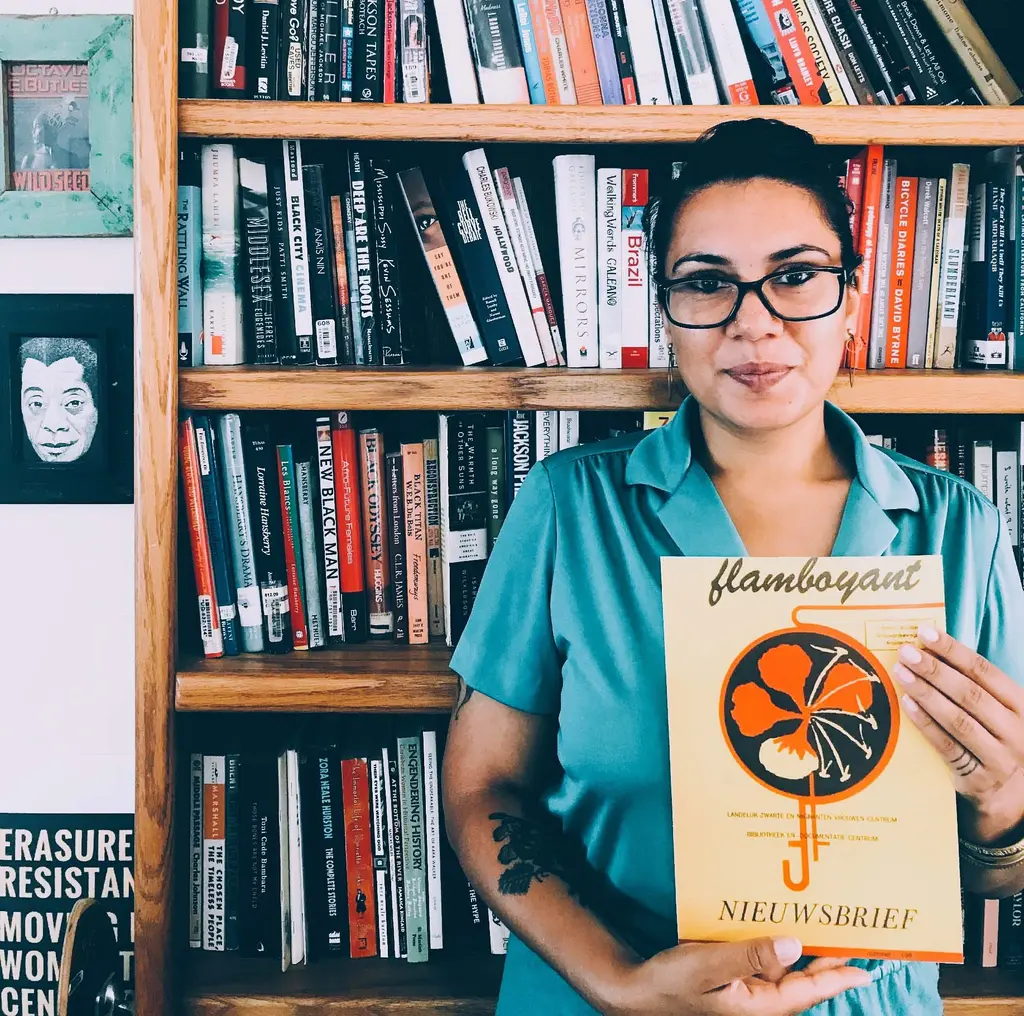 Woman with black glasses and blue dress stands in front of bookcase with magazine Flamboyant in her hands