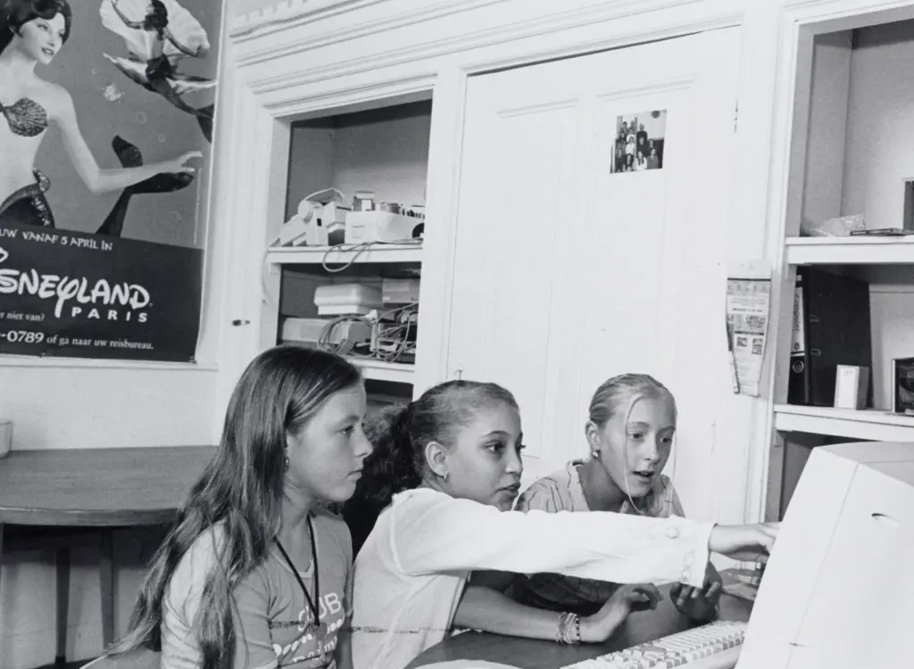 Drie meiden zitten achter desktopcomputer uit 2000 met op de achtergrond een reclameposter voor Disneyland met een zeemeermin