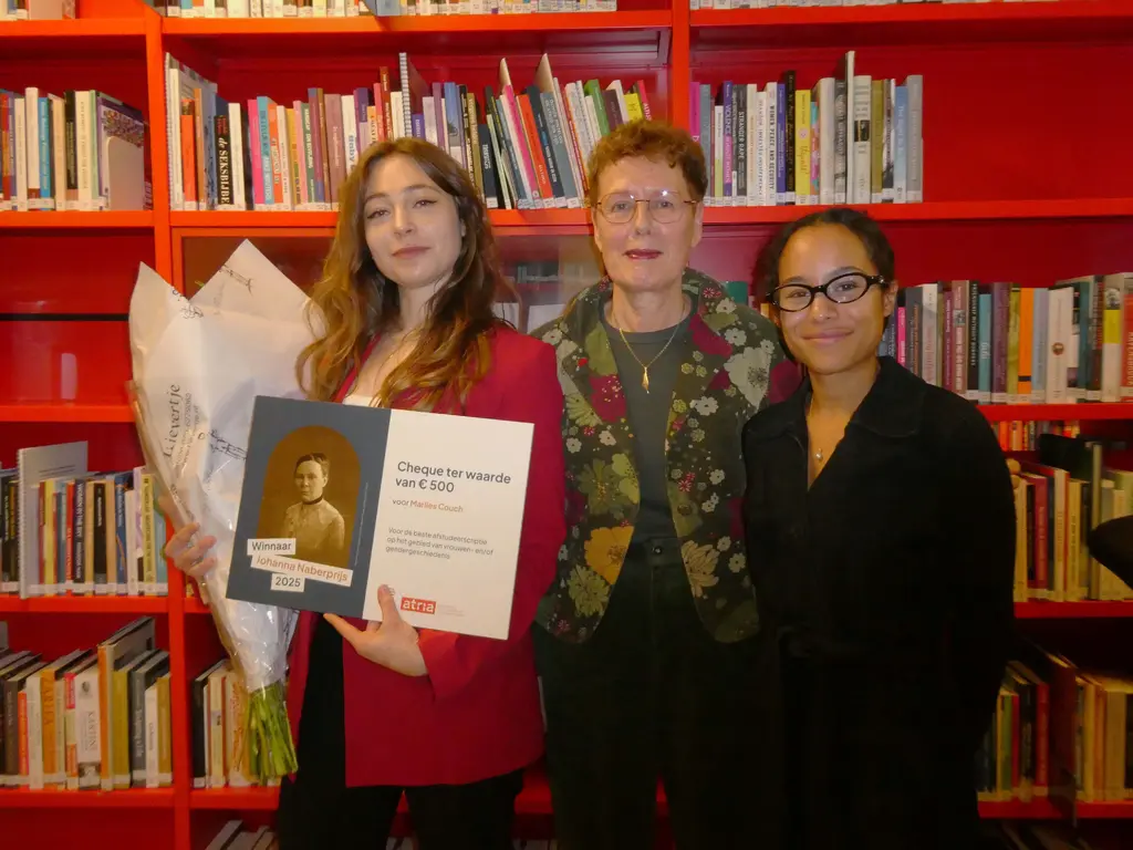 Marlies Couch with flowers and large cheque worth 500 euros, Francisca de Haan, and Alex Erica Mary McQueen, standing in front of red bookcases