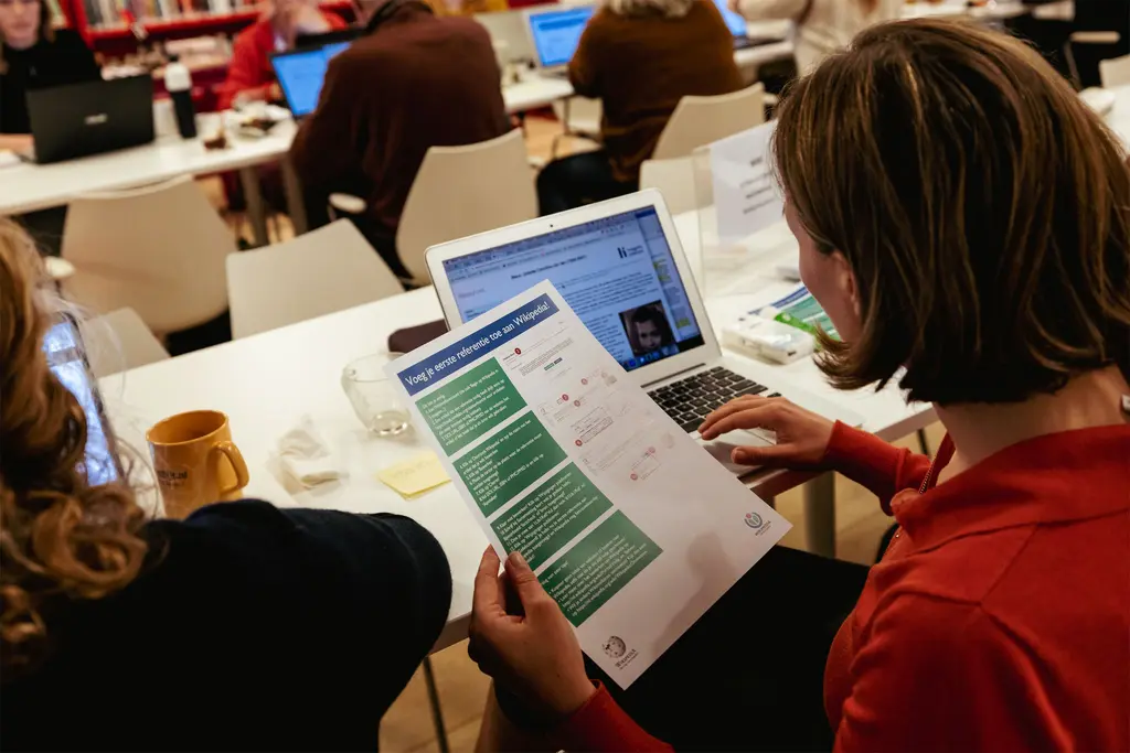 Vrouw zit achter laptop met instructie hoe je een referentie moet invoegen op Wikipedia in haar linkerhand