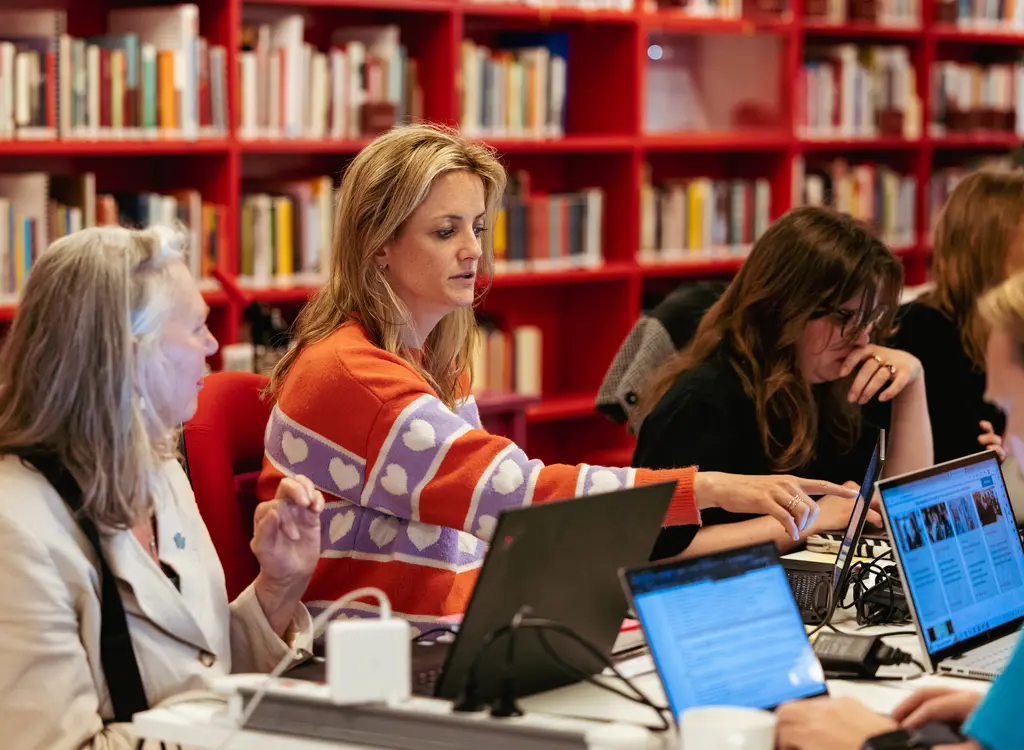 Groep mensen zitten aan tafel achter laptops te werken en 2 vrouwen overleggen met elkaar