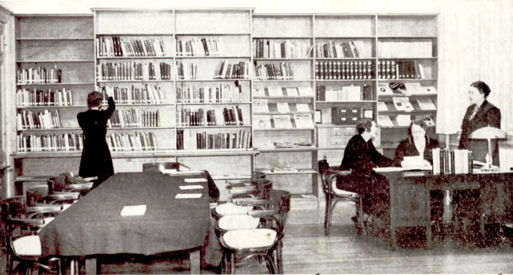 Ruimte met grote tafel en bureau waaraan twee vrouwen zitten en een vrouw staat met op de achtergrond een aantal hoge boekenkasten met vrouw die boek pakt