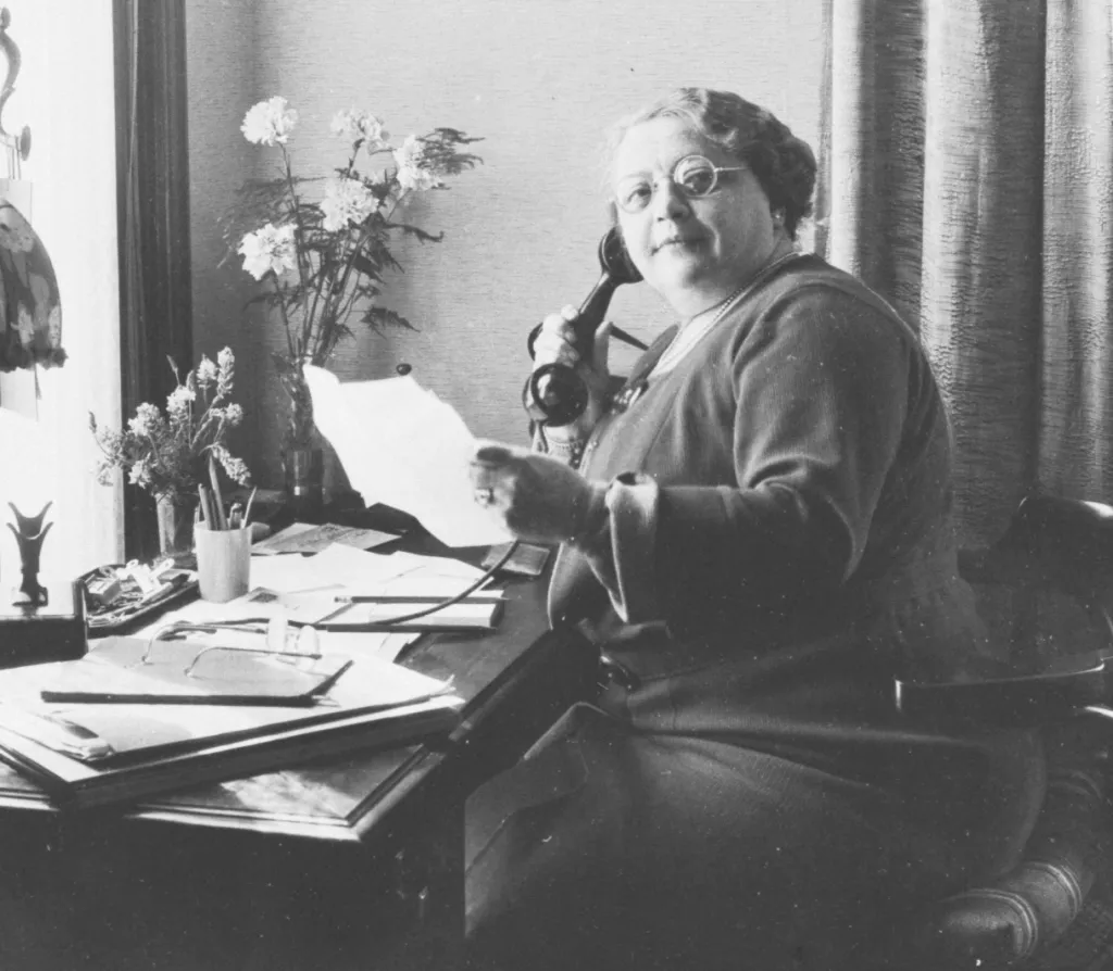 Rosa Manus zit aan een bureau met een telefoon in haar hand. Om haar heen liggen stapels met brieven en twee vasen met bloemen.