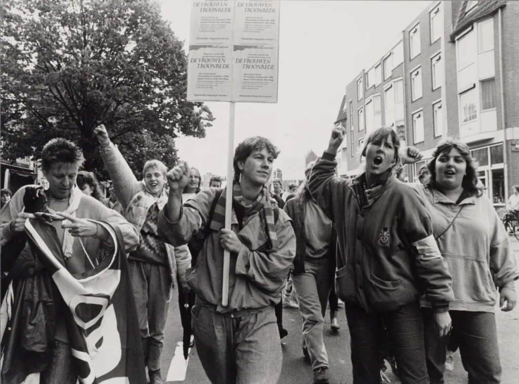 jonge vrouwen lopen op straat te protesteren en een van hen houdt een bord omhoog met tekst De Vrouwentroonrede